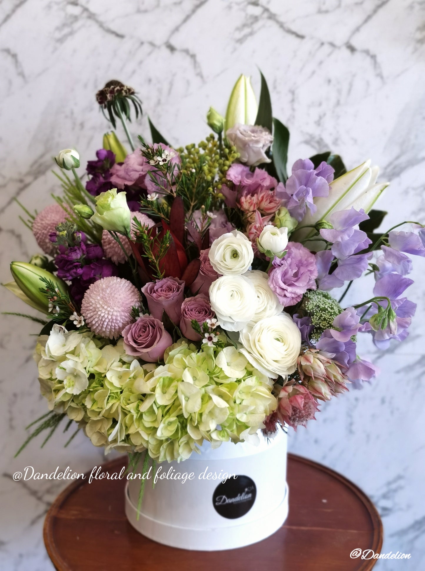 A round premium flat hat box arranged with a variety of flowers in shades of purple and white.