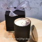 A contemporary black glass jar soy candle with a silver lid, decorated with a purple bow and placed next to a black box with a purple flower design.
