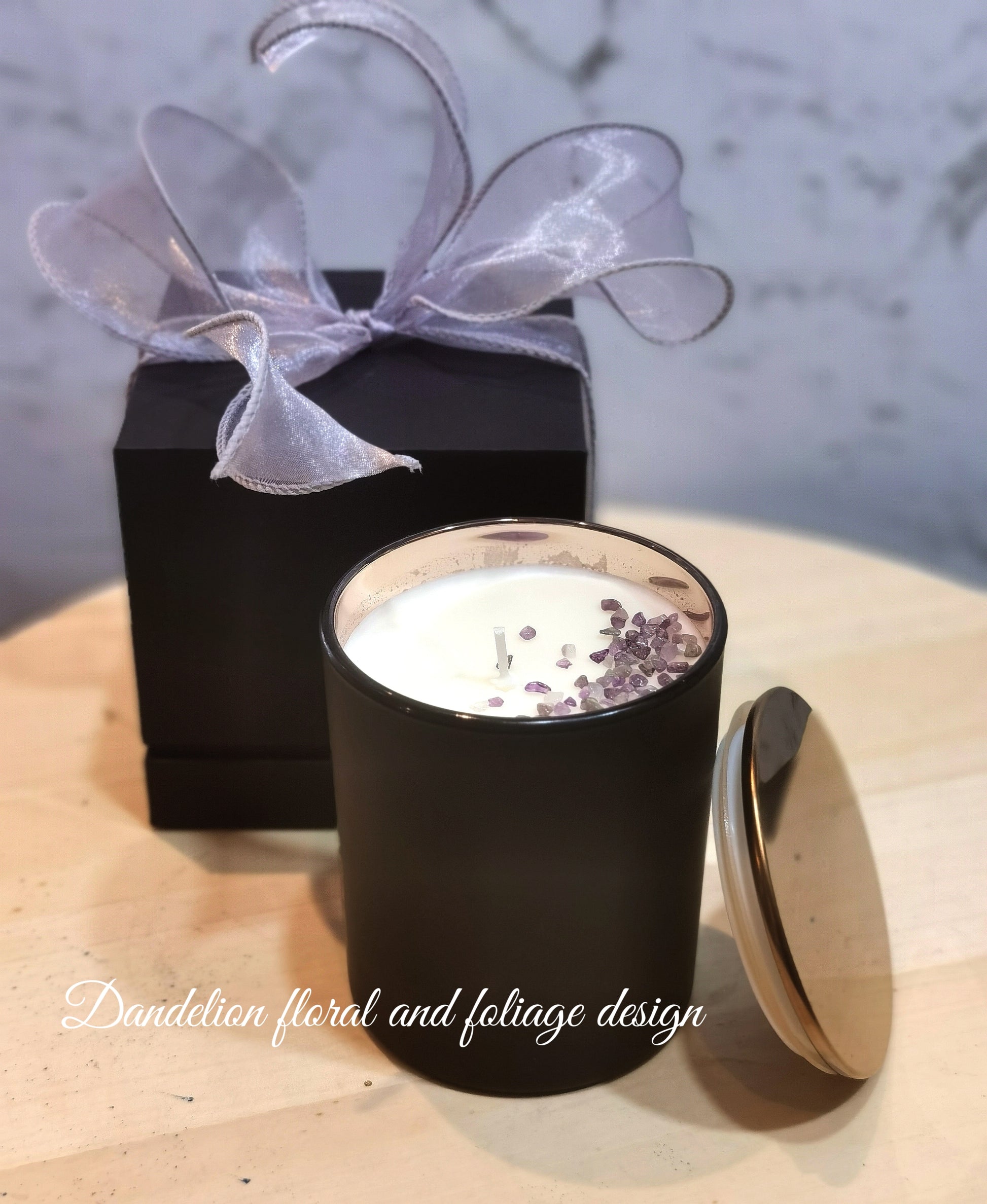 A contemporary black glass jar soy candle with a silver lid, decorated with a purple bow and placed next to a black box with a purple flower design.