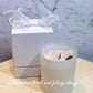 A contemporary white glass jar soy candle with a white lid, decorated with a white bow and placed next to a whitebox with a pink flower design.