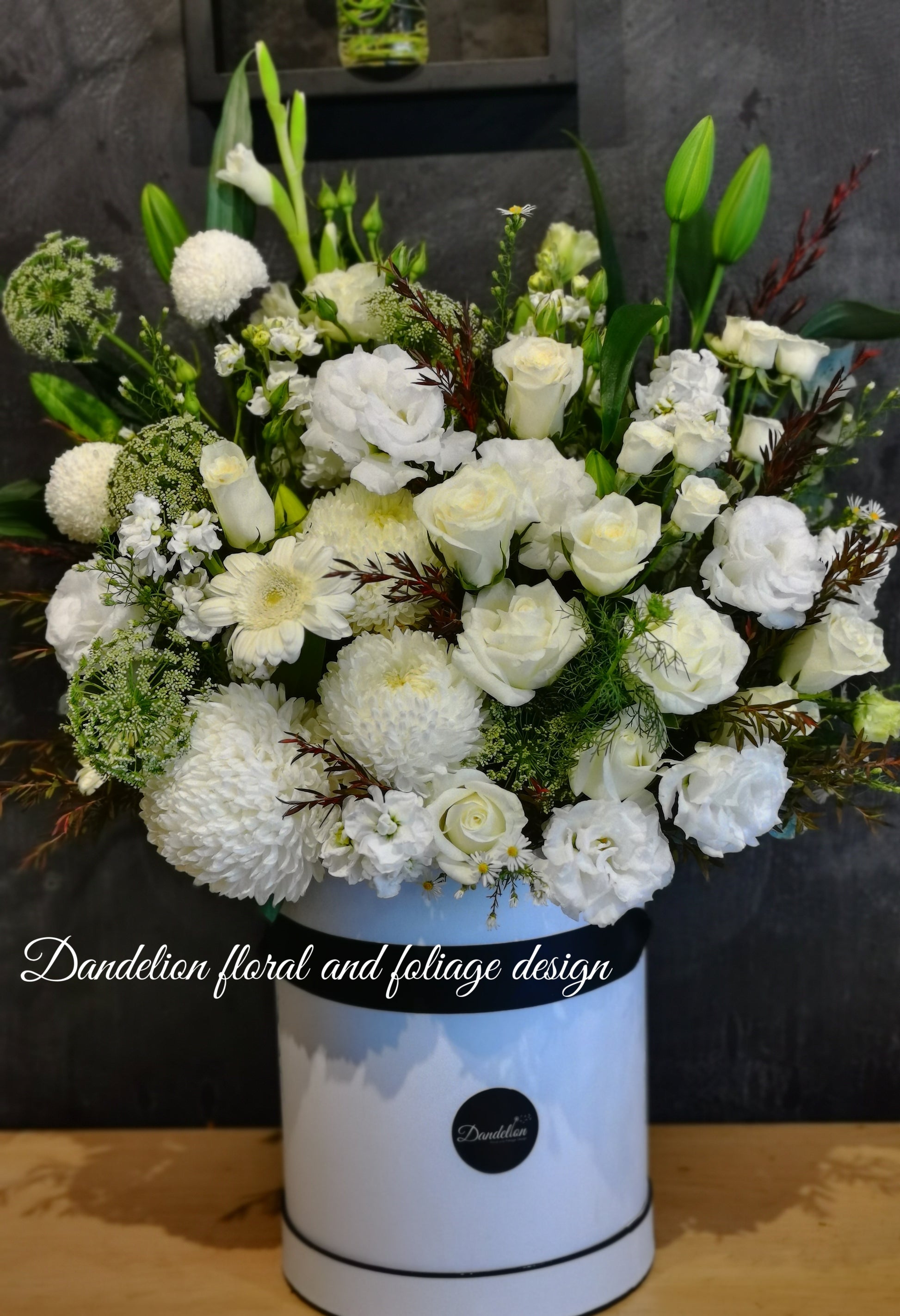 A round tall white hat box filled with white and green flowers, arranged white classic yet in a modern shape. Wedding, engagement or a sad occasion, this arrangement will fulfill what you are in need for.