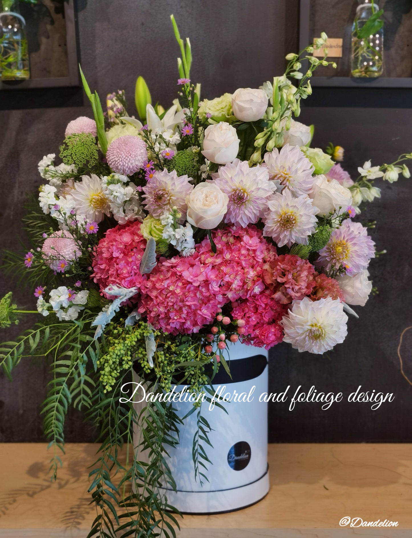 A large pink hat box flower arrangement featuring a variety of beautiful and premium flowers, displayed in a shopfront setting. 
