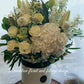A floral arrangement in a hat box with a variety of flowers including Hydrangea, Peony roses, and Oriental lilies, presented in shades of pink, white, and green. 
