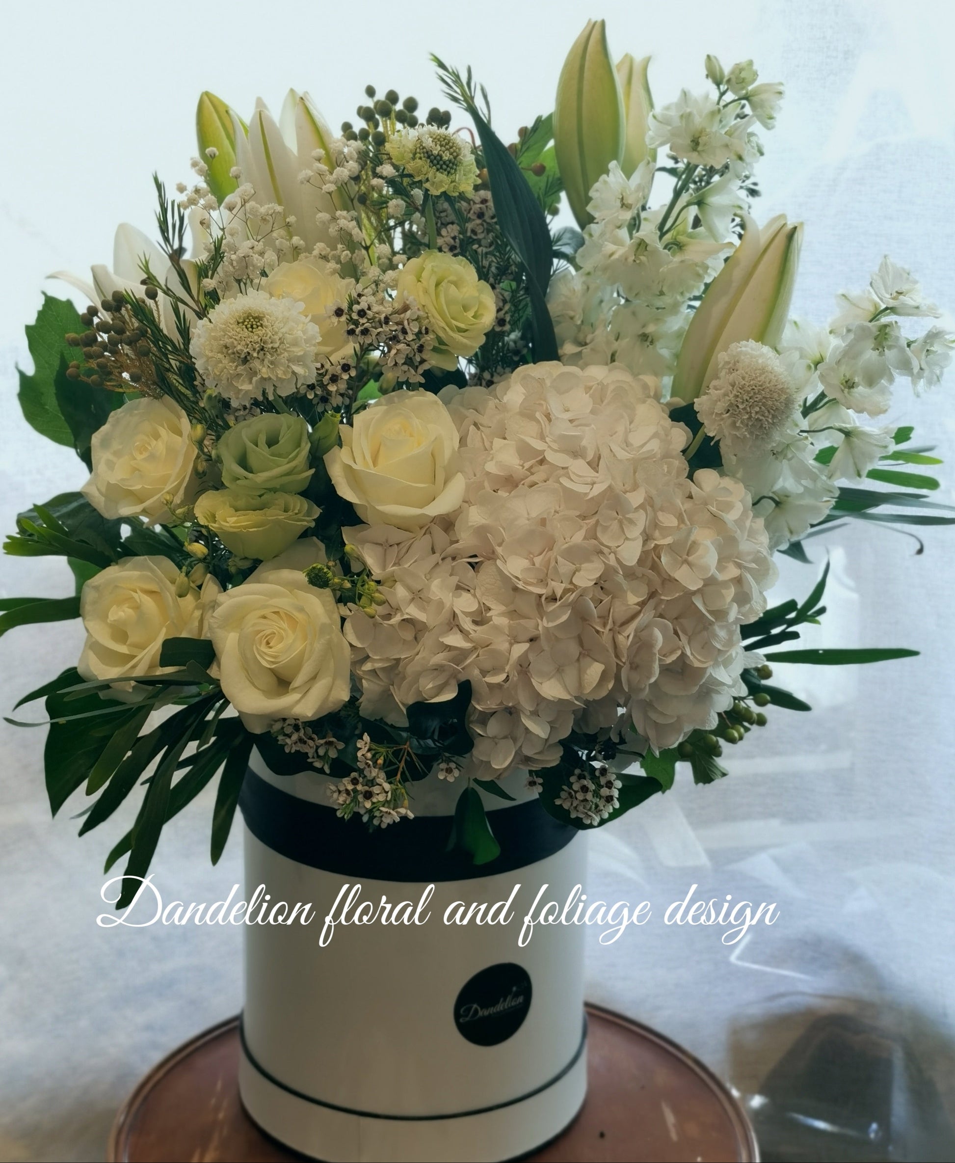 A floral arrangement in a hat box with a variety of flowers including Hydrangea, Peony roses, and Oriental lilies, presented in shades of pink, white, and green. 