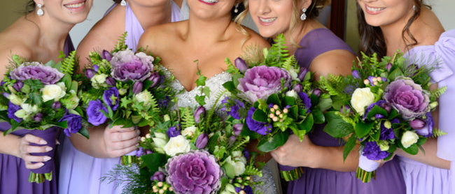 Five women in purple dresses holding floral bouquets indoors.