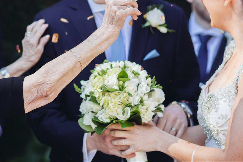 Wedding ceremony with a woman holding a bouquet and an elderly woman using a fan, surrounded by guests outdoors.