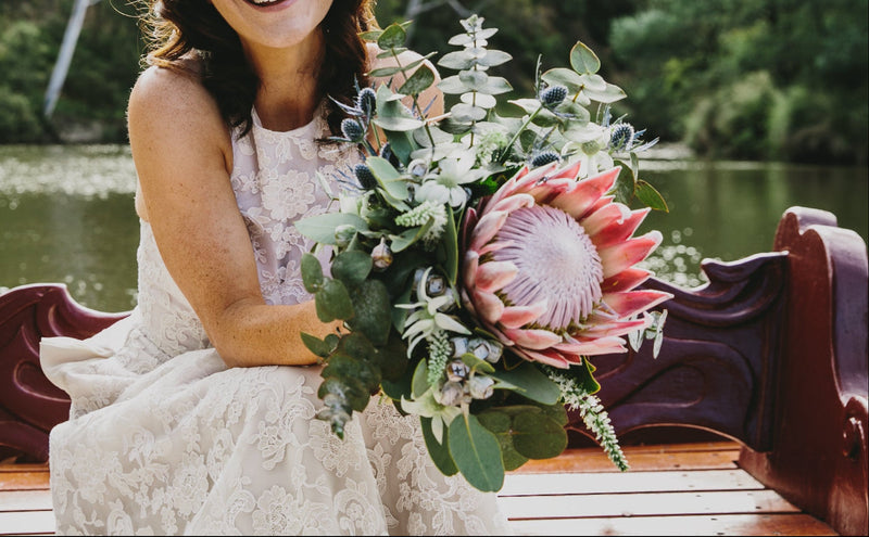 Woman in a wedding dress holding a bouquet by a lake with greenery
