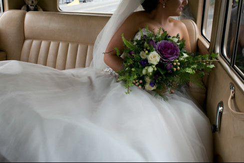 Bride in a wedding dress with a bouquet, sitting in a car with a man in a suit.
