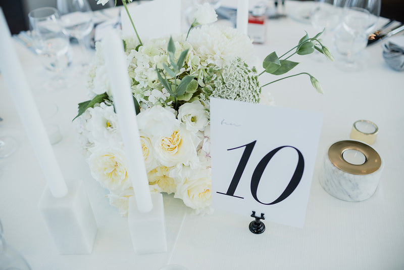 Table setting with white flowers, a table number card, and candles on a white tablecloth for lush wedding