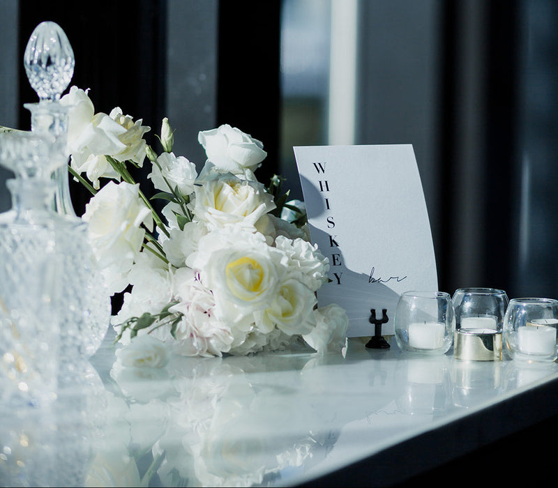Decorative table setting with white flowers, candles, and a card on a reflective surface for luxury wedding