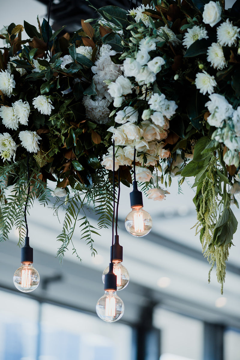 Decorative white and green arrangement of flowers and hanging light bulbs against a blurred indoor background