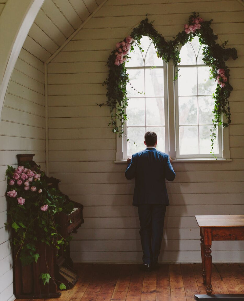 Man standing in a small room with wooden floor and white walls, looking out of a window decorated with floral garlands.