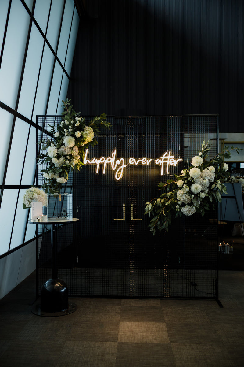 Neon sign reading 'happily ever after' with white and green floral arrangements on a dark background