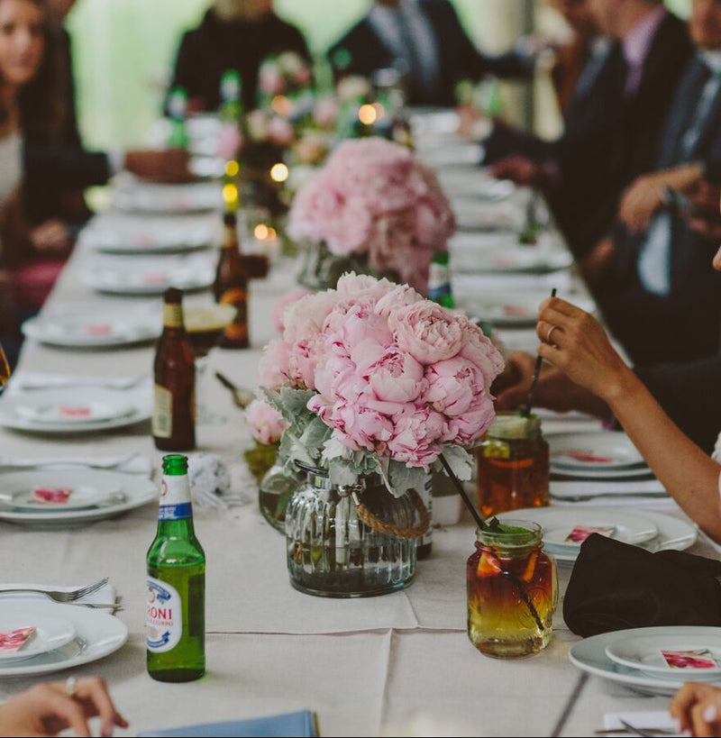 Long dining table set for a formal event with pink floral centerpieces, people seated around, and bottles on the table.