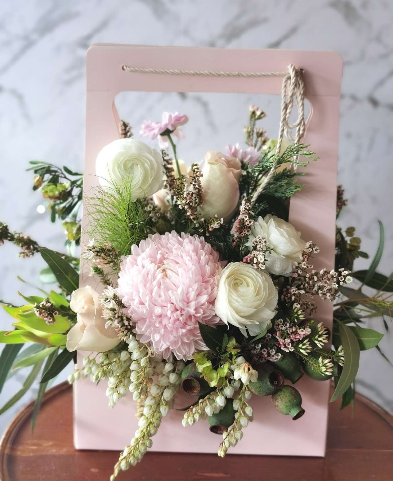A floral carry box arrangement with a variety of flowers including a pink carnation, white roses, and green foliage, presented in a rectangular pink box.