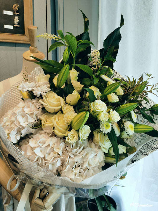 A stunning floral arrangement featuring a mix of white roses, lilies and other seasonal fresh flowers with accompanying green foliage. 