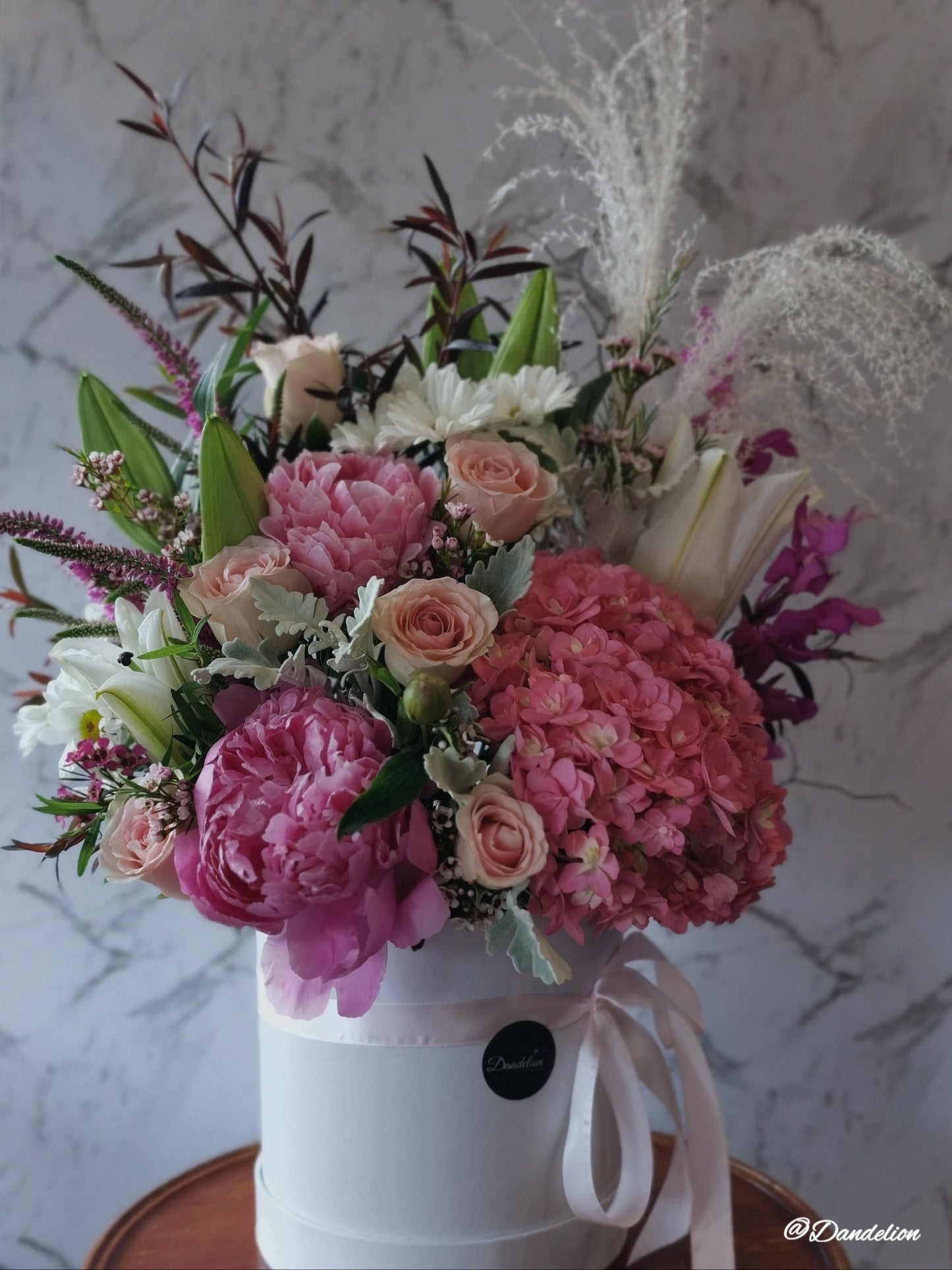A floral arrangement in a hat box with a variety of flowers including Hydrangea, Peony roses, and Oriental lilies, presented in shades of pink, white, and green. 