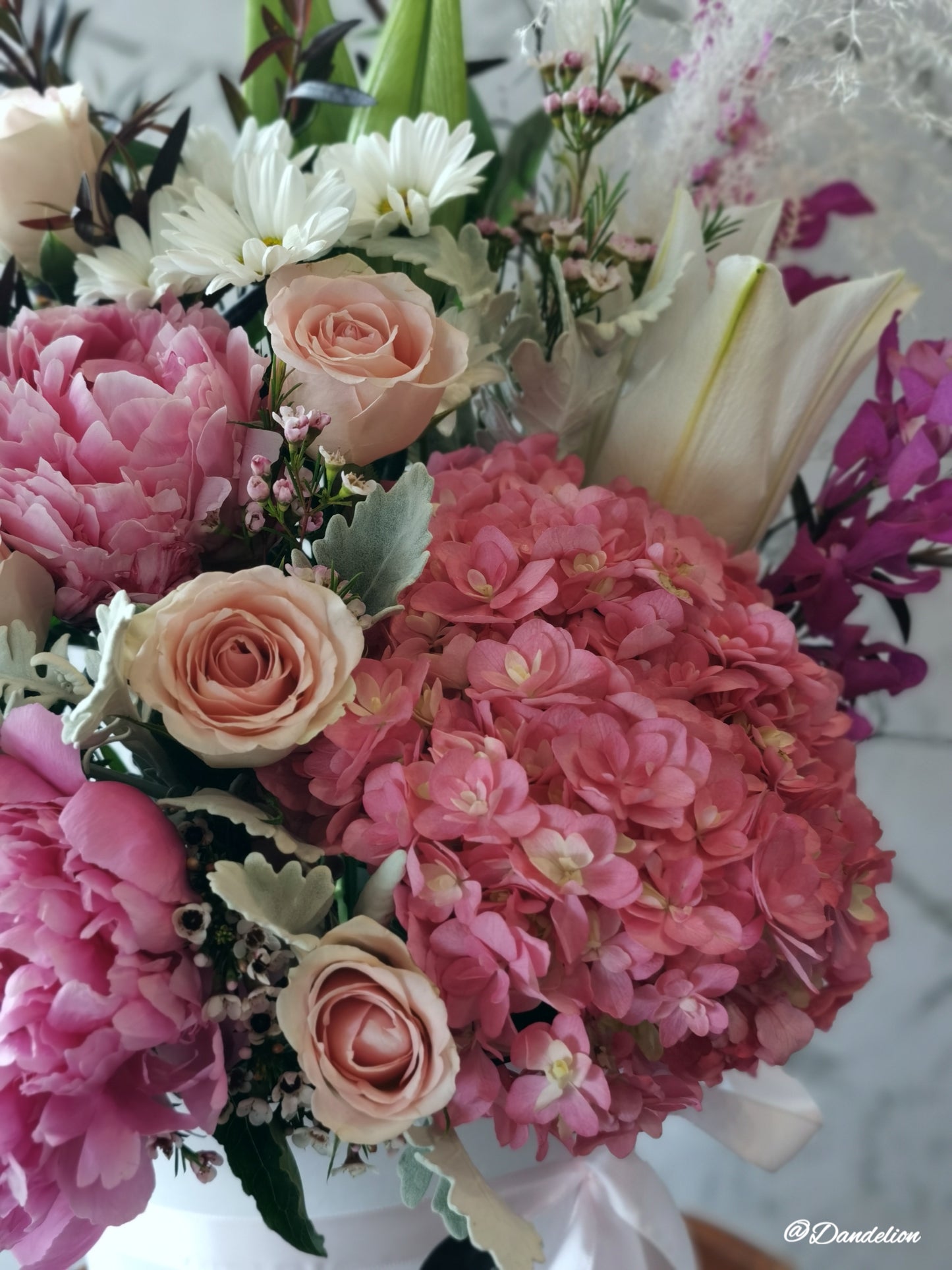A floral arrangement in a hat box with a variety of flowers including Hydrangea, Peony roses, and Oriental lilies, presented in shades of pink, white, and green. 
